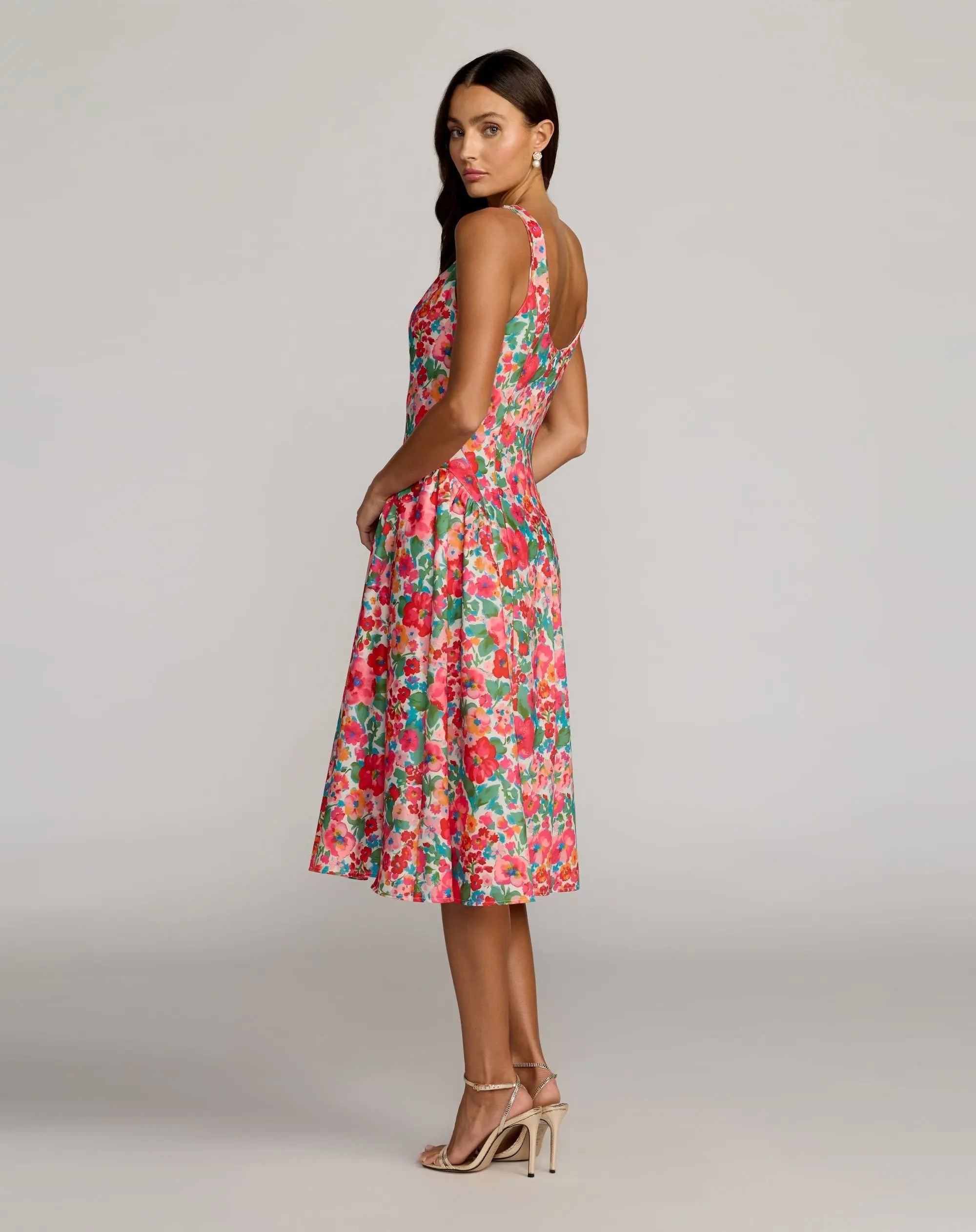 A woman wearing the Mac Duggal 37155 Floral Midi Wedding Guest Dress, featuring a sleeveless square neckline and floral print crepe fabric, paired with strappy high heels, stands in a studio with a neutral background.